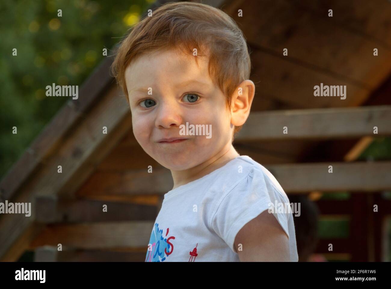 Portrait d'un adorable garçon à tête rouge et yeux bleus, portant un t-shirt blanc dans une aire de jeux par temps ensoleillé Banque D'Images