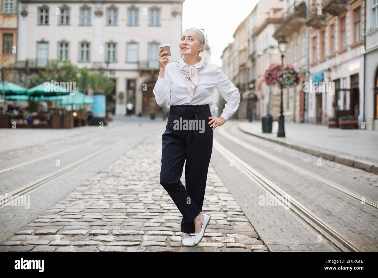 Femme positive dans une tenue elegante posant dans la rue avec une tasse de cafe dans les mains. Bonne femme retraite qui passe du temps libre activement a l exterieur Photo Stock