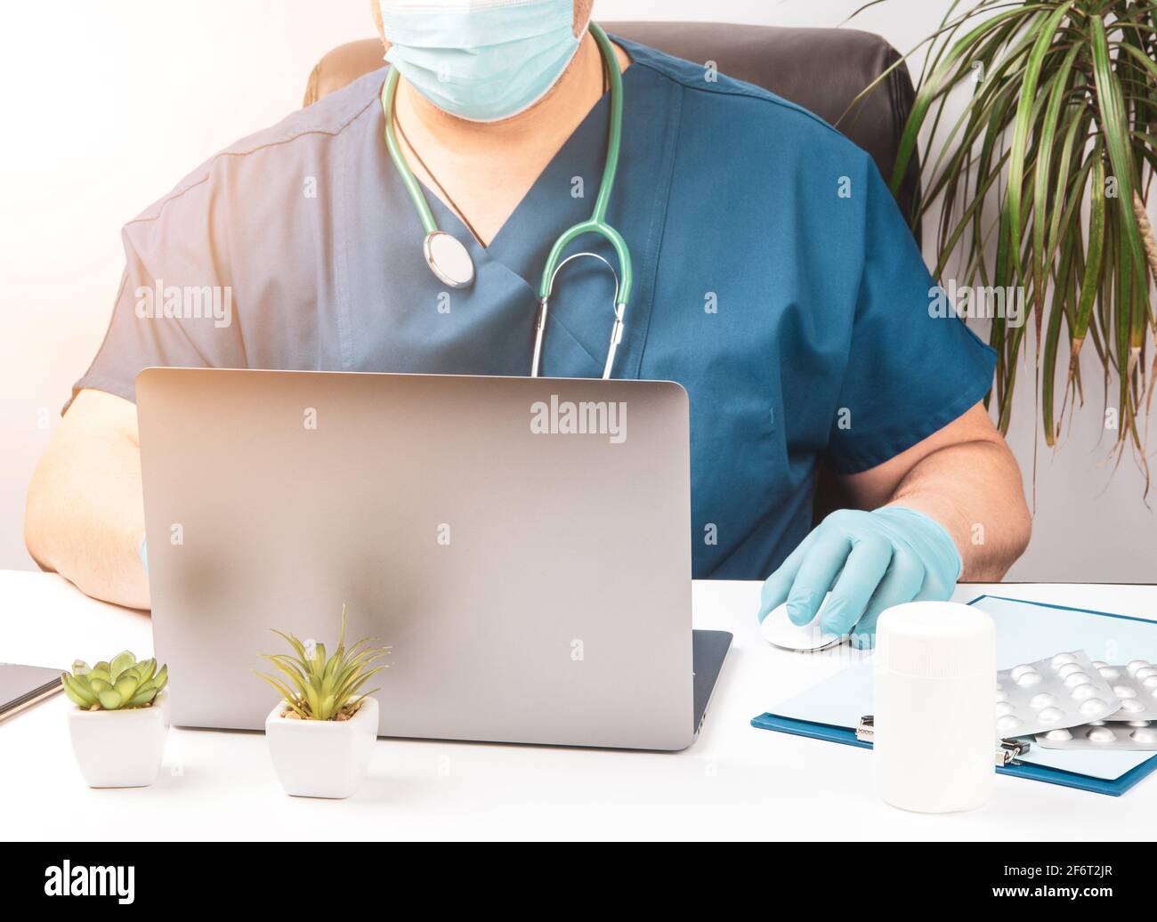 homme médecin assis à une table blanche en uniforme bleu et gants en latex, spécialiste travaille sur un ordinateur portable, concept de télémaintenance, thérapeute Banque D'Images