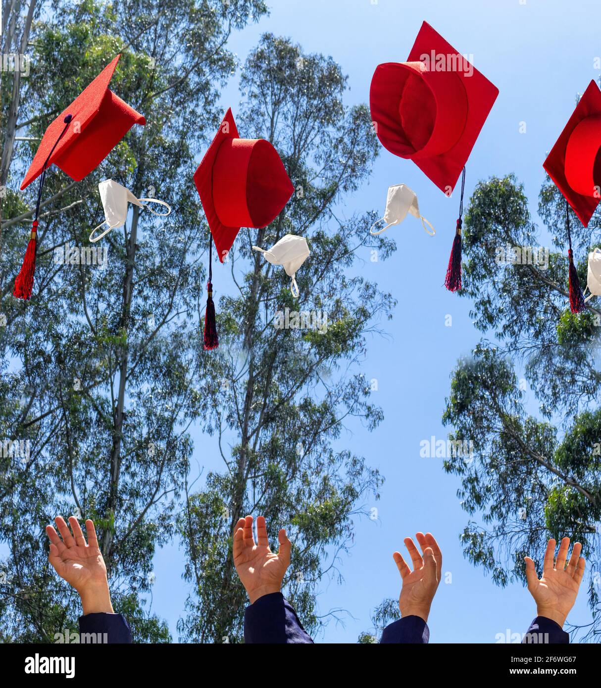 Les mains des élèves qui lancent des chapeaux et des masques de remise des diplômes rouges dans l'air au-dessus d'un ciel bleu . Graduation et distanciation sociale Banque D'Images