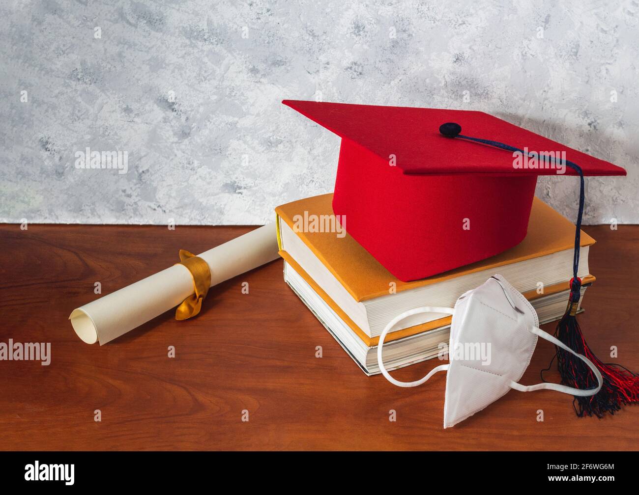 Encore la vie avec un chapeau de graduation rouge sur quelques livres à côté d'un diplôme et un masque sur un bureau en bois. Concept de la graduation et de la distanciation sociale Banque D'Images