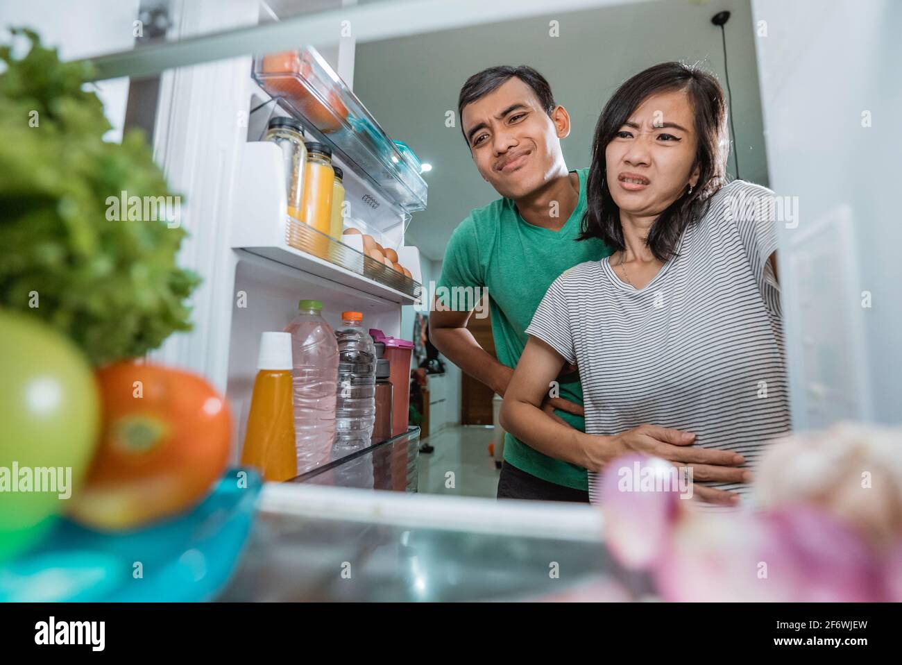 un couple asiatique tient son ventre tout en ouvrant la porte du réfrigérateur à la recherche de quelque chose à manger Banque D'Images