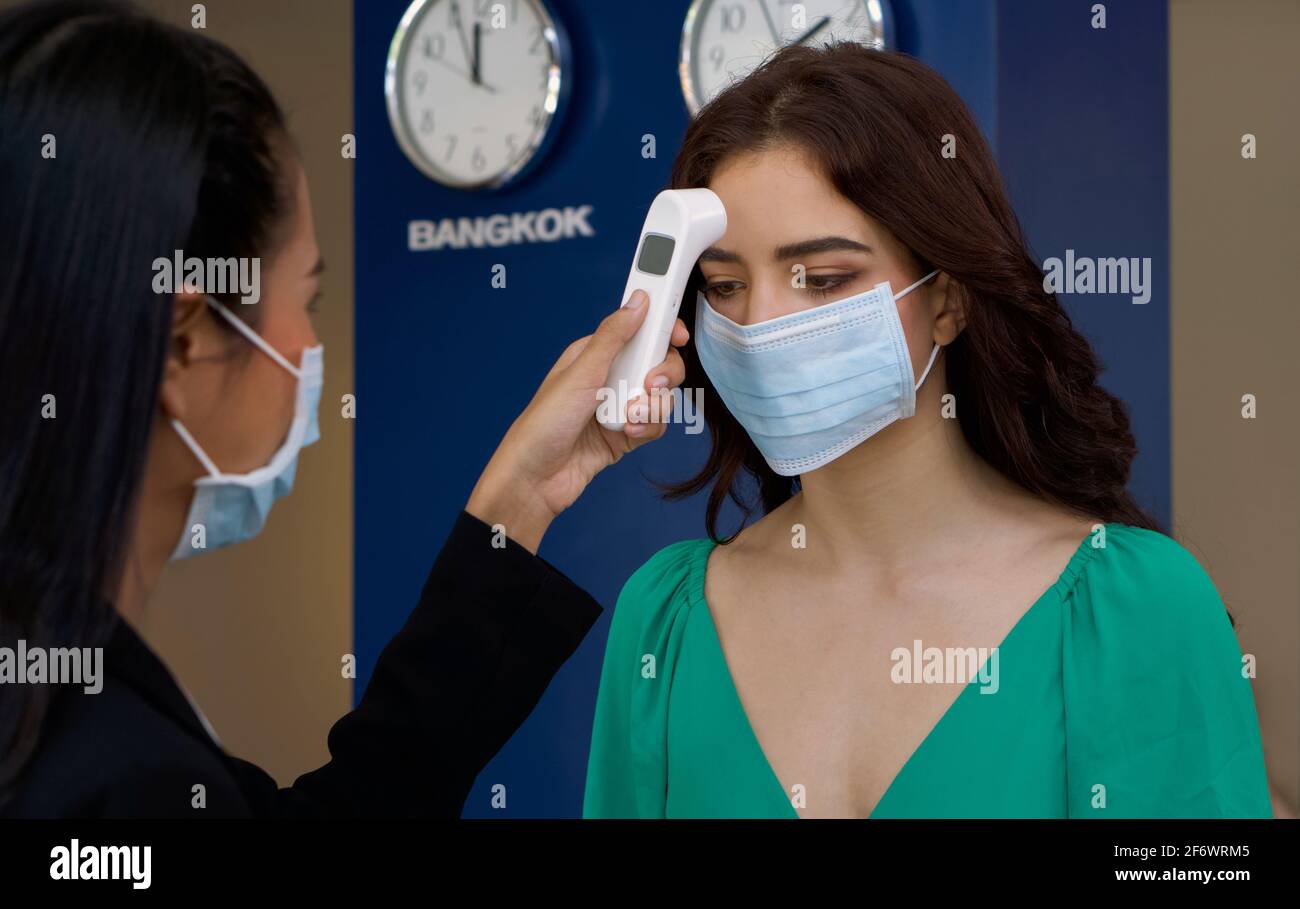 Touriste caucasien dans le clothe vert porter un masque lors de la mesure de la température avec un outil de diagnostic de surveillance de la fièvre. Le mur du fond a une horloge à dire Banque D'Images