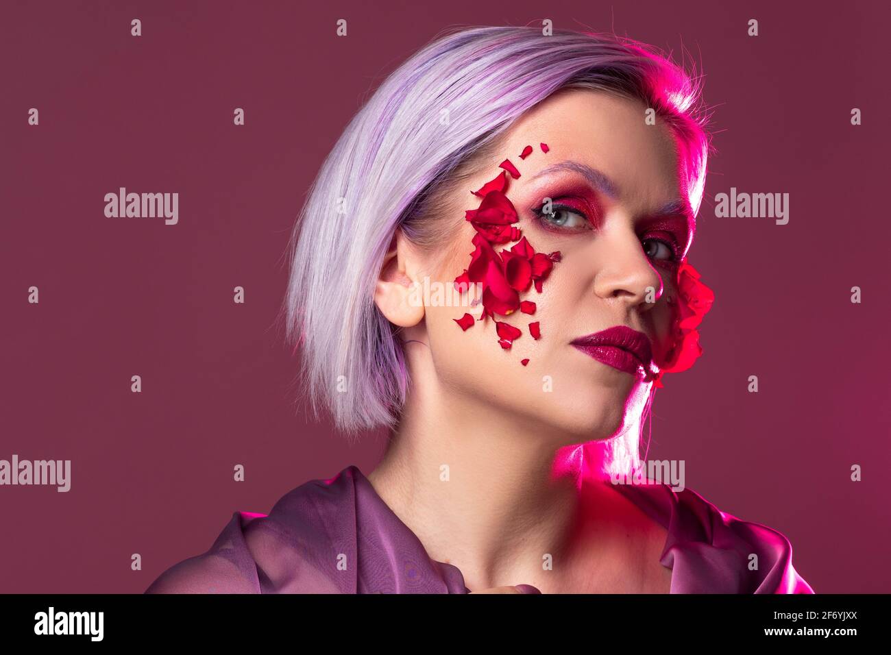 Jeune femme élégante et lumineuse avec un maquillage coloré et des pétales de fleur sur son visage, une image de mode sur un fond lilas. Banque D'Images