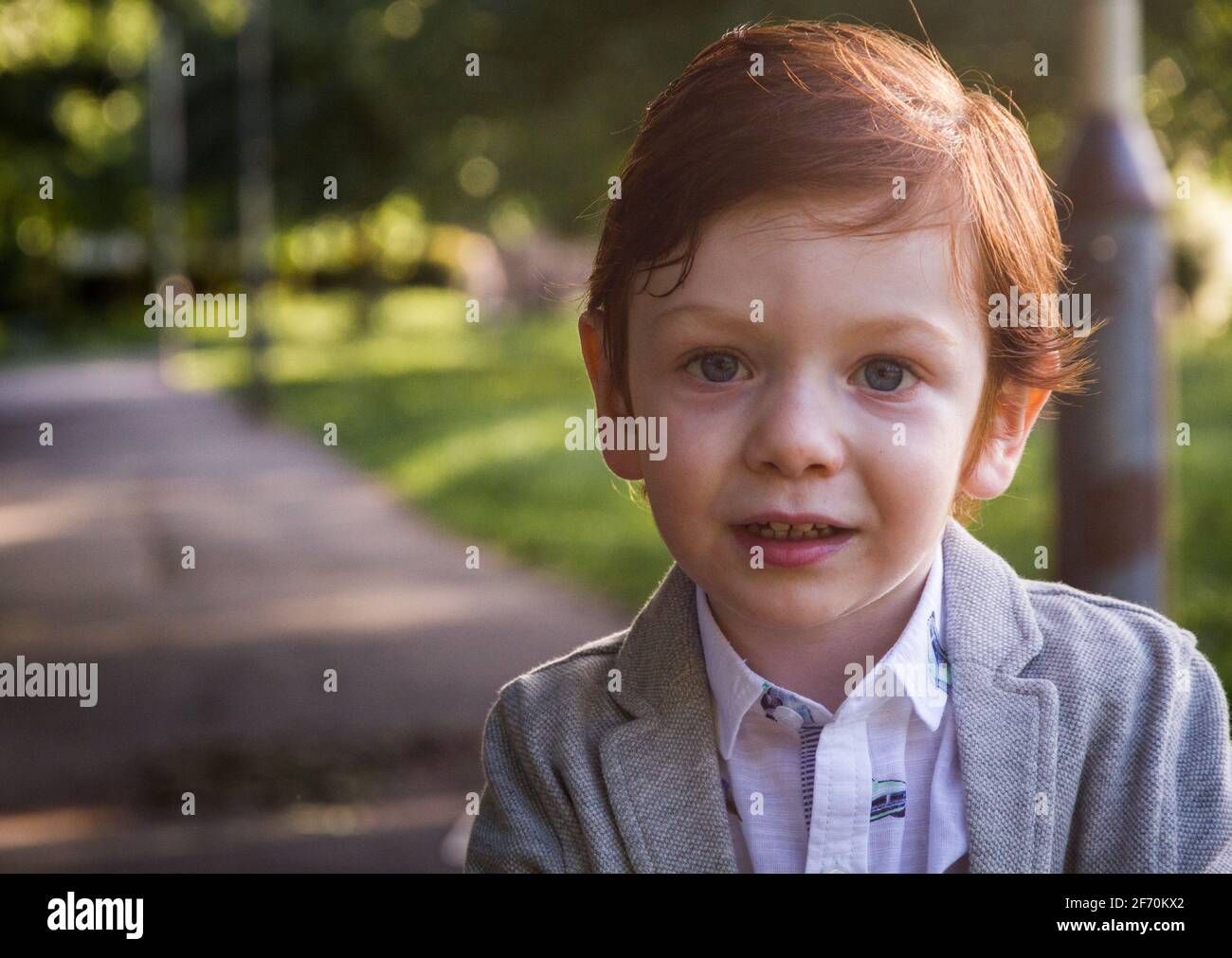 Portrait d'un adorable garçon à tête rouge et yeux bleus, portant un blazer gris et une chemise blanche dans un parc par temps ensoleillé Banque D'Images