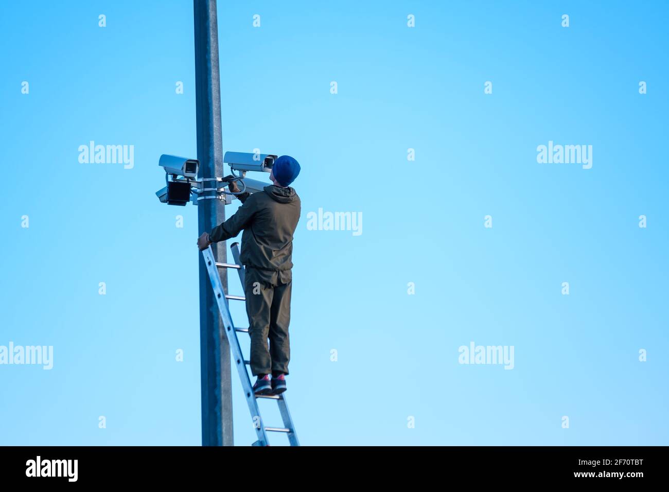 Vue arrière d'un travailleur masculin méconnaissable debout sur une échelle et installation de caméras de surveillance modernes sur un poteau contre un ciel bleu sans nuages Banque D'Images