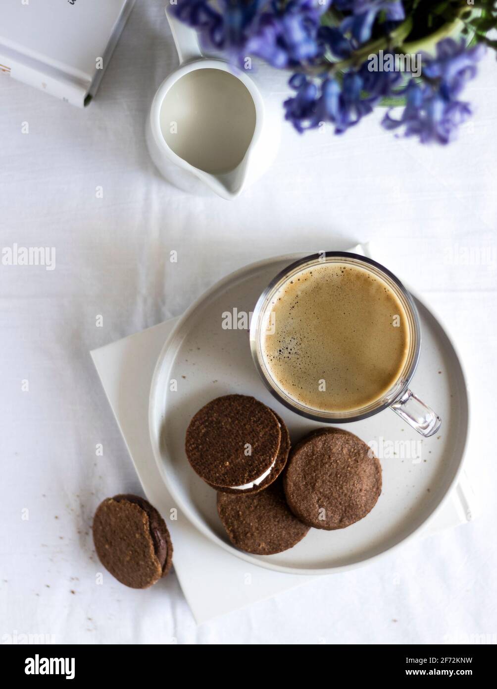 Pause café du matin - café fraîchement préparé servi avec quelques biscuits au chocolat végétalien sur un livre, un pot à lait et des fleurs en arrière-plan. Banque D'Images