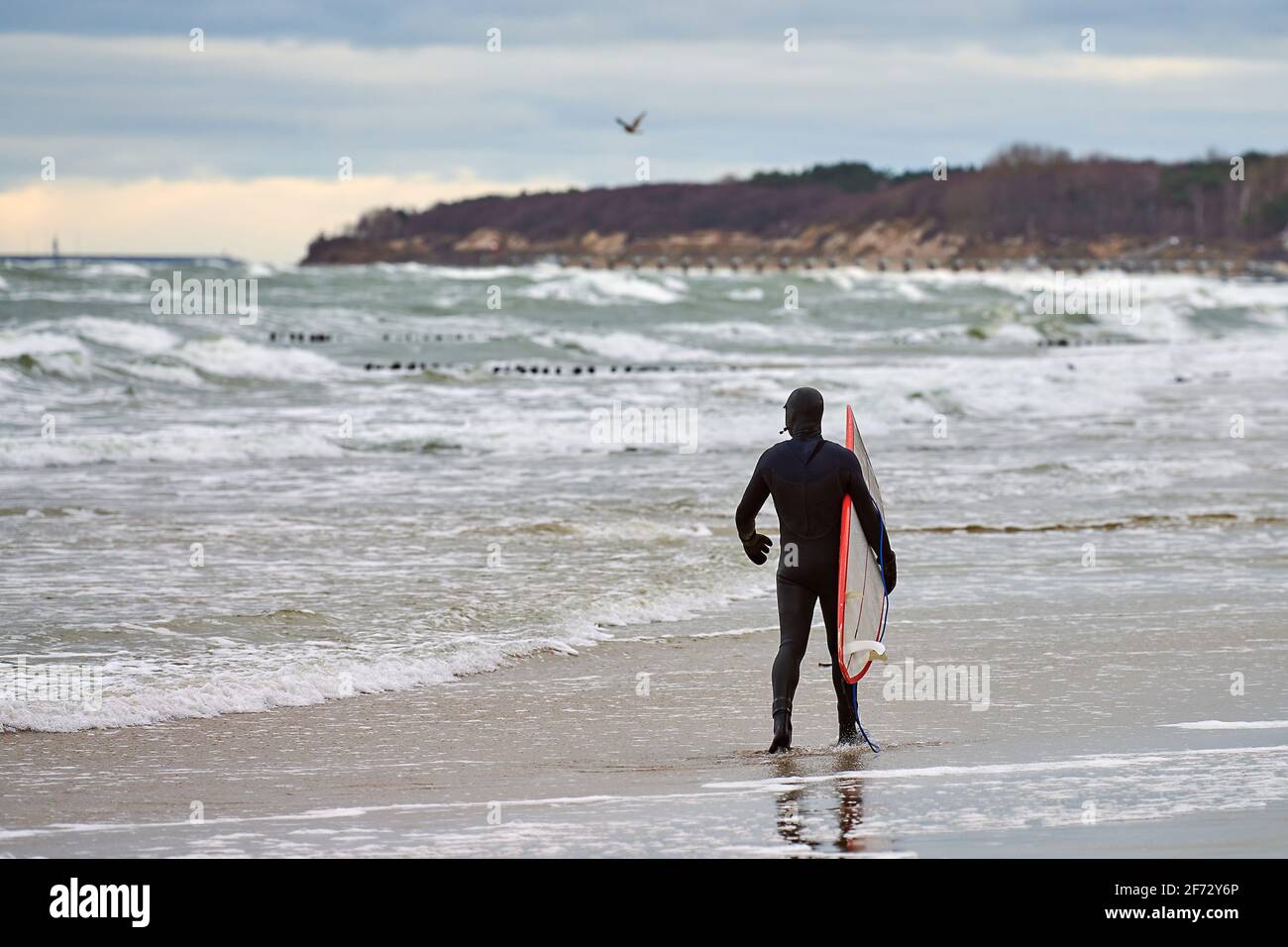 Surfeur masculin en maillot de bain noir marchant le long de la mer et tenant le surf blanc dans sa main. Journée chaude, beau ciel nuageux, scène de la nature. Sport et outdo Banque D'Images