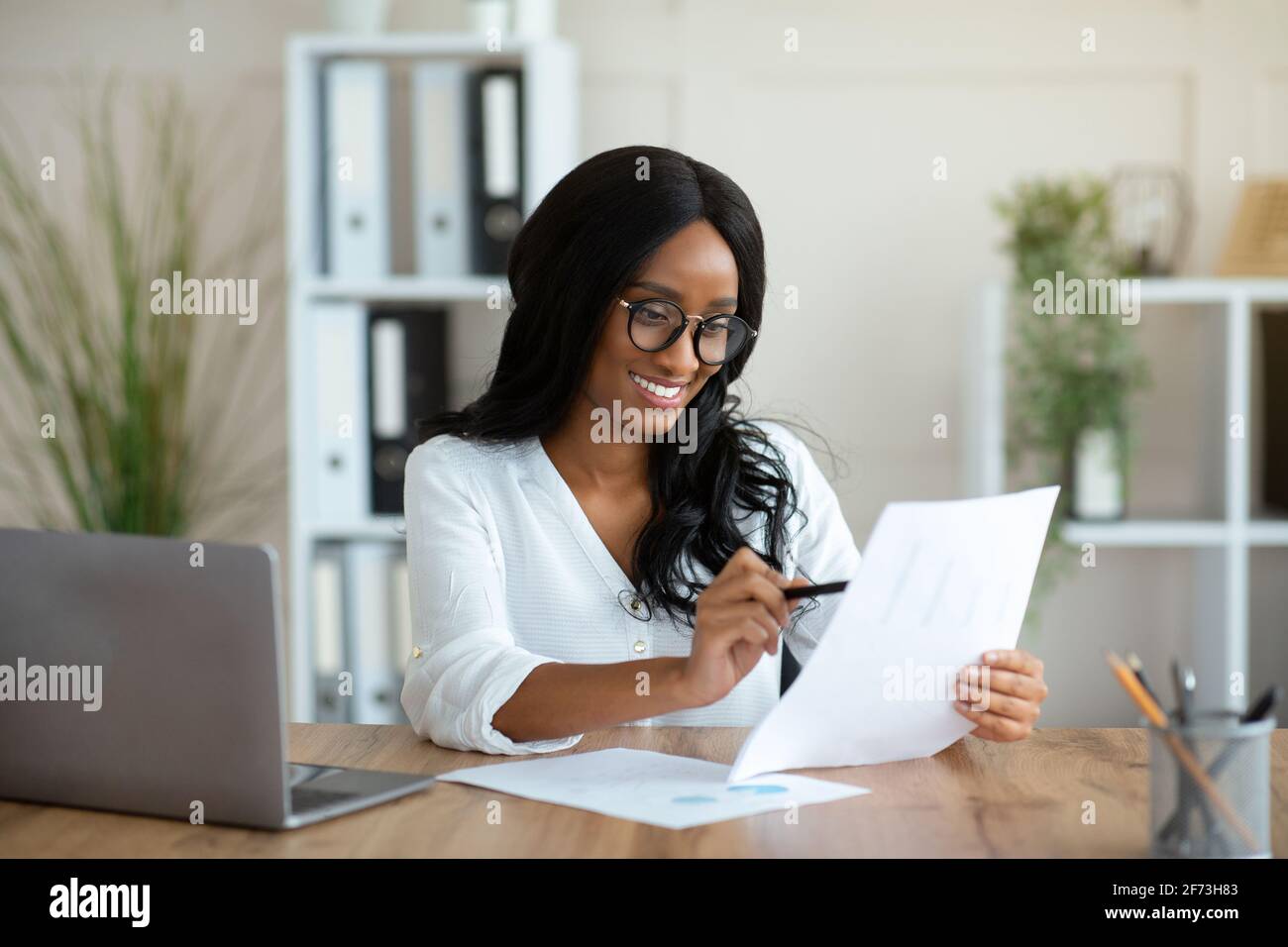 Femme d'affaires noire attrayante regardant à travers le document à son bureau dans bureau Banque D'Images