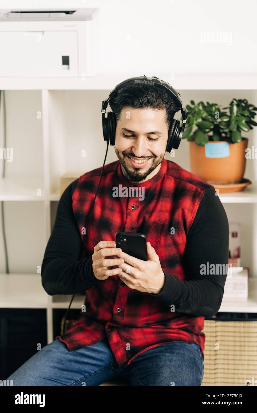 Musicien masculin barbu adulte dans un casque de messagerie texte sur un téléphone portable dans la salle de séjour Banque D'Images