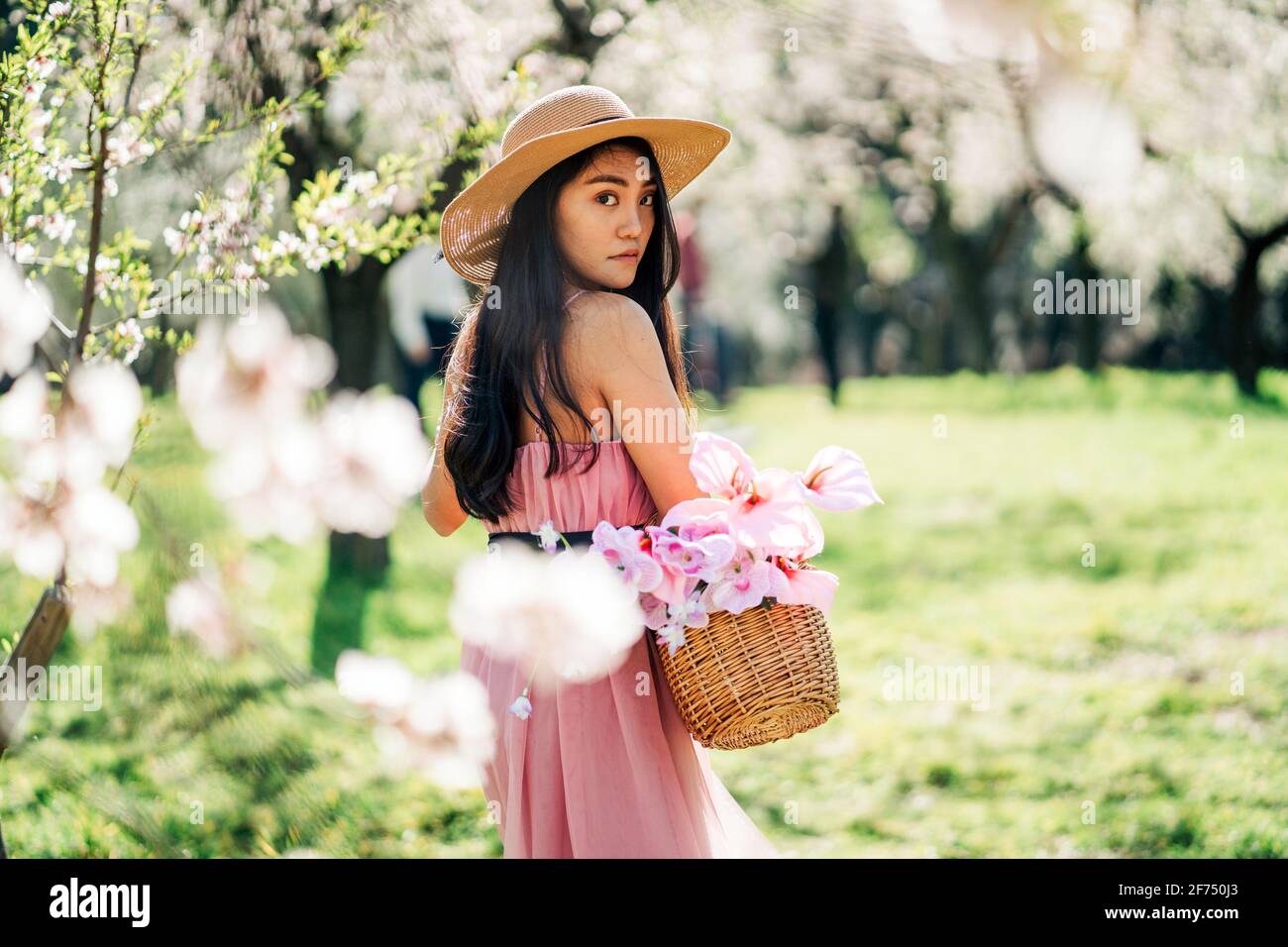 Vue arrière de la femme en robe et chapeau de paille debout avec panier dans le jardin en fleur en regardant par-dessus l'épaule vers appareil photo Banque D'Images