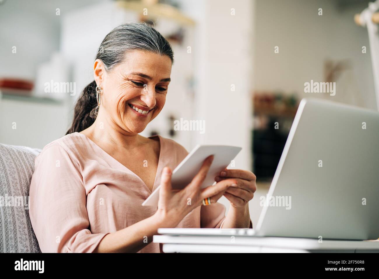 Femme d'âge moyen et gaie assise sur un canapé près d'un netbook moderne tout en regardant la photo dans un appartement moderne avec cuisine arrière-plan flou Banque D'Images