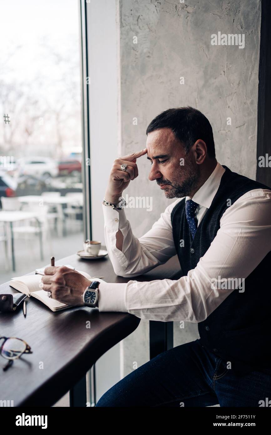 Vue latérale d'un homme d'affaires hispanique mature et pensif touchant le front à la table avec l'ordre du jour dans la cafétéria Banque D'Images