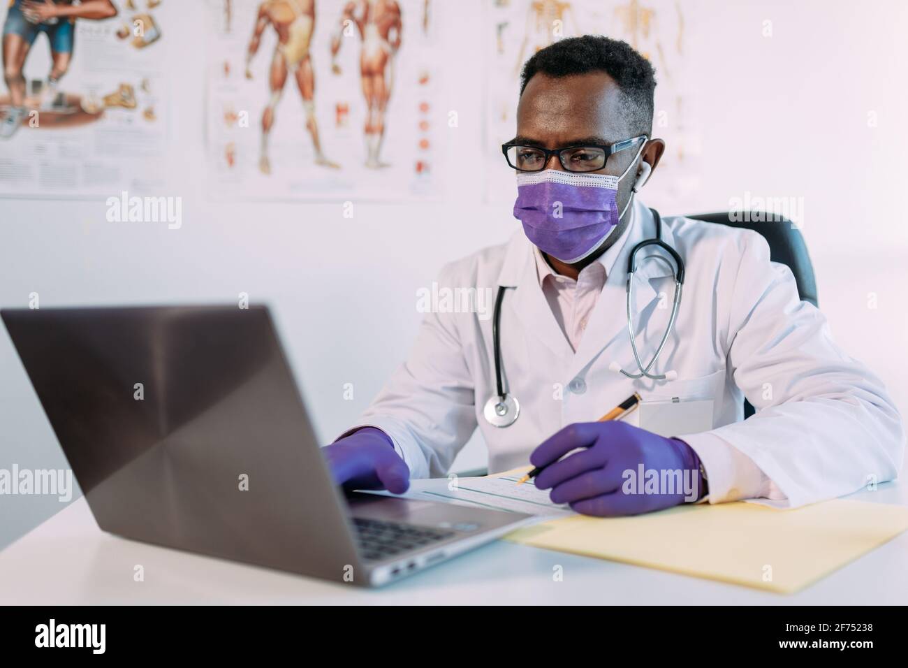 Médecin afro-américain en lunettes de vue travaillant avec le patient en ligne sur netbook lors de l'écriture dans le dossier patient à la table de l'hôpital Banque D'Images
