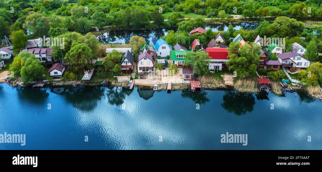 Petites maisons suburbaines sur la rive de la rivière avec le ciel bleu reflet dans l'eau et arbres verts forêt sur fond, vue aérienne de drone. Banque D'Images