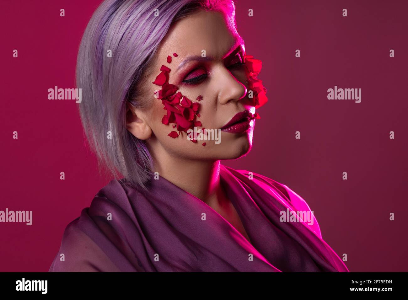 Jeune femme élégante et lumineuse avec un maquillage coloré et des pétales de fleur sur son visage, une image de mode sur un fond lilas. Banque D'Images
