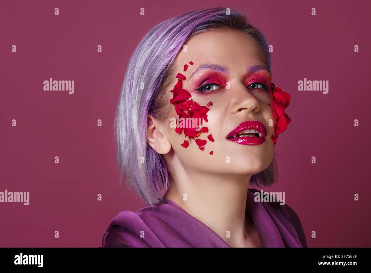 Jeune femme élégante et lumineuse avec un maquillage coloré et des pétales de fleur sur son visage, une image de mode sur un fond lilas. Banque D'Images