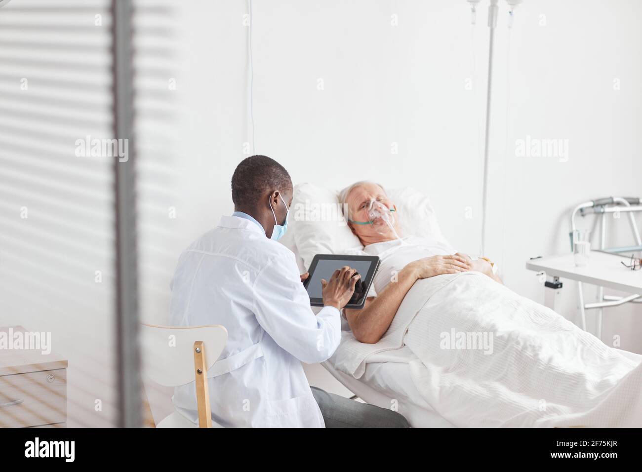 Vue arrière portrait d'un médecin afro-américain utilisant une tablette numérique tout en étant assis par un patient âgé dans le lit d'hôpital, espace de copie Banque D'Images