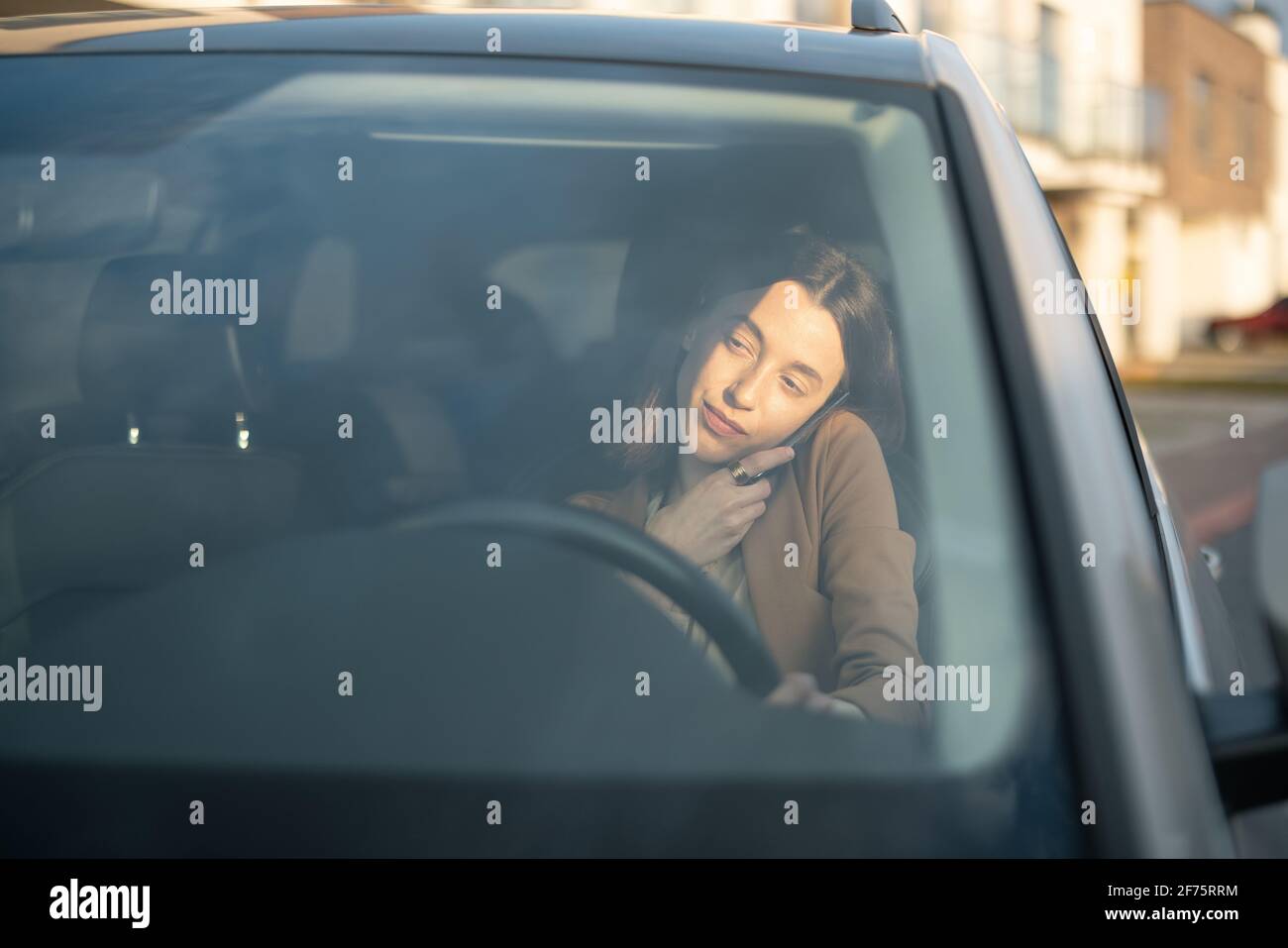 Une jeune femme parle au téléphone tout en conduisant une voiture et en tenant un téléphone sur son épaule. Femme d'affaires qui va travailler de la maison. Vue par le pare-brise. Banque D'Images