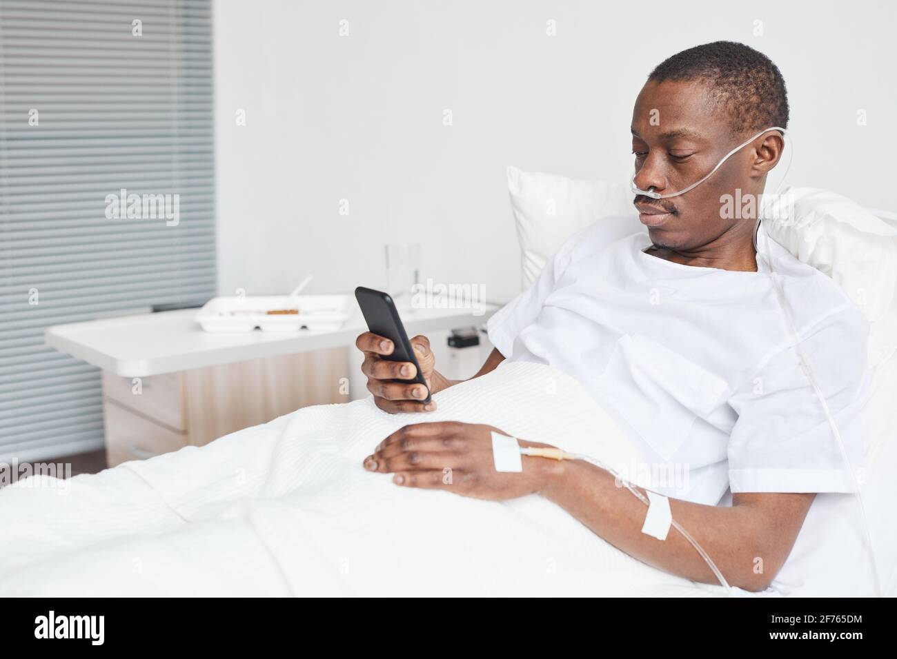 Portrait de l'homme afro-américain dans le lit d'hôpital et en utilisant un smartphone avec des tubes de supplémentation en oxygène, espace de copie Banque D'Images