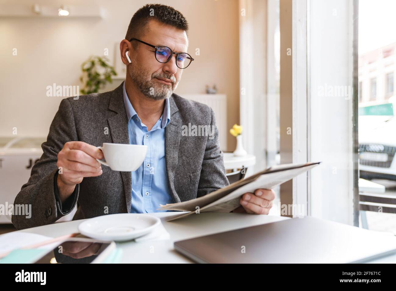 Homme d'affaires charmant et confiant lisant des journaux et buvant du café dans un café à l'intérieur, en portant des écouteurs Banque D'Images