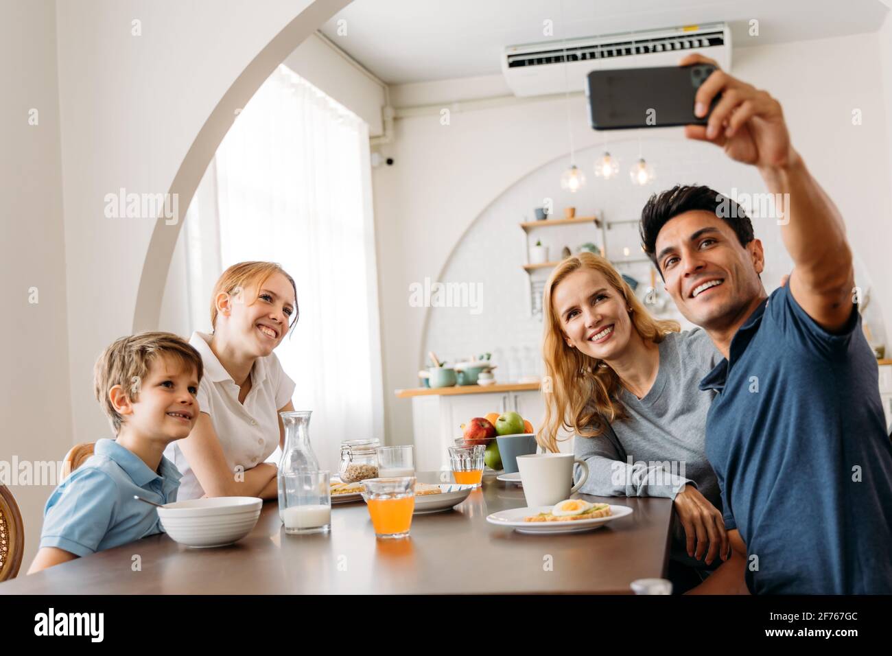 Groupe de quatre familles caucasiennes prenant un selfie en prenant un petit déjeuner sur une table de salle à manger. Père, mère, fille et fils appréciant le temps heureux avec la cuisine en arrière-plan. Banque D'Images
