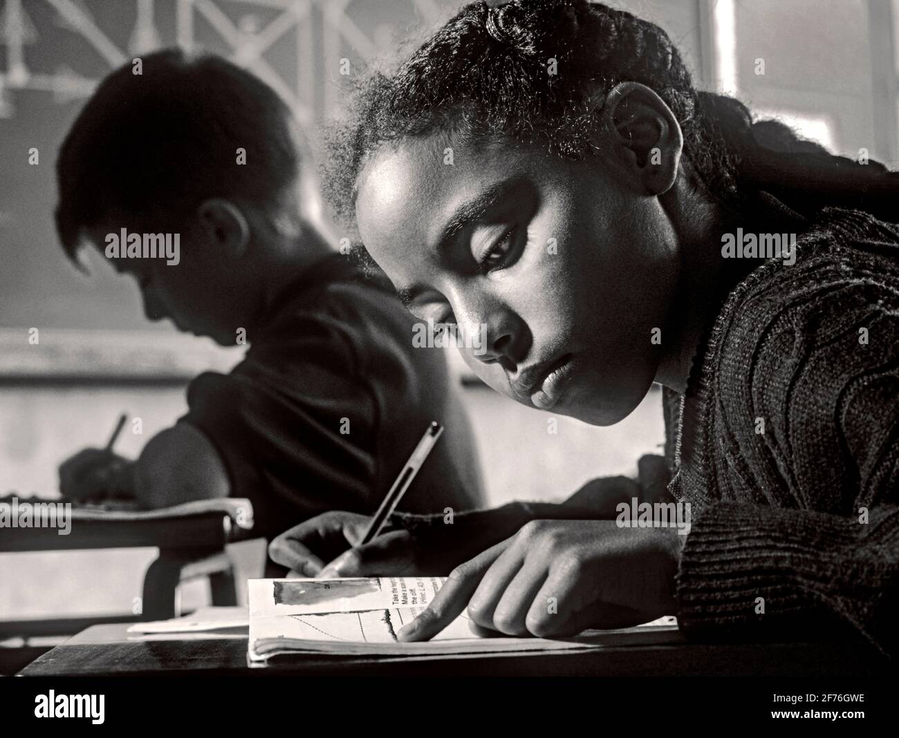 Junior fille bureau de classe écrire école scolaire noir africain 9-11 des années en classe écrivant à son bureau avec un garçon blond Et la géométrie de l'école projettent un tableau noir derrière l'image B&W. Banque D'Images