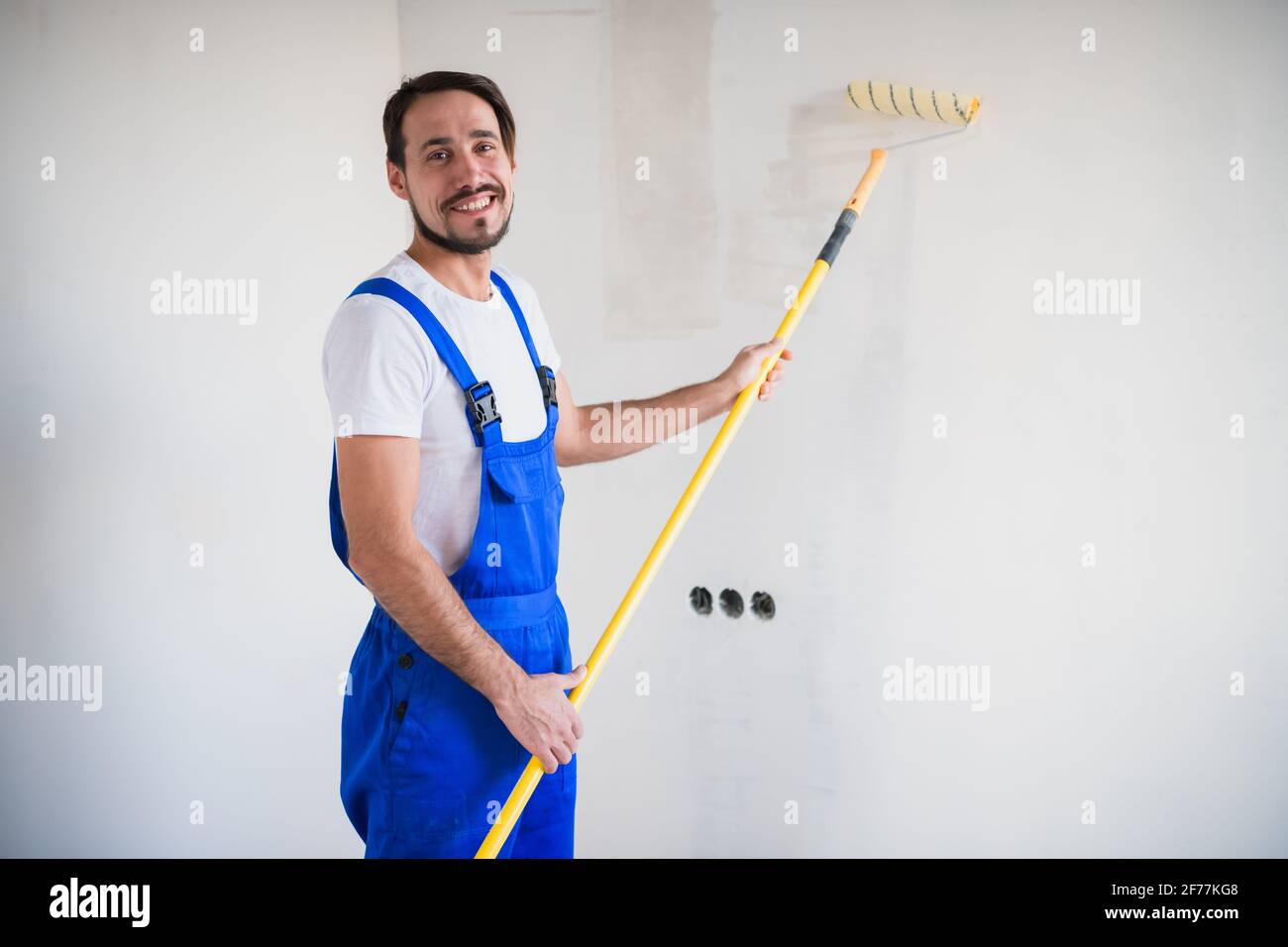 Un peintre barbu en vêtements de travail peint le mur avec un rouleau. Il sourit amical et pose pour la caméra Banque D'Images