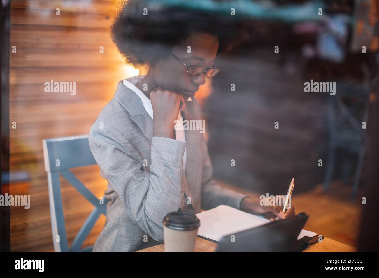 Jeune femme africaine concentrée en costume d'affaires écrivant des notes tout en étant assise à table de café avec une tablette numérique. Concept de travail à distance, personnel et technologie. Banque D'Images
