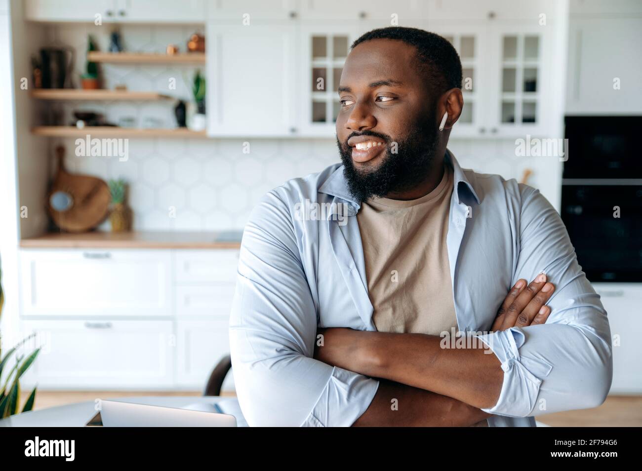 Portrait d'un jeune homme afro-américain plein de joie et confiant avec une barbe portant un casque debout dans le salon, portant des vêtements élégants, regardant sur le côté, bras croisés devant lui, souriant Banque D'Images