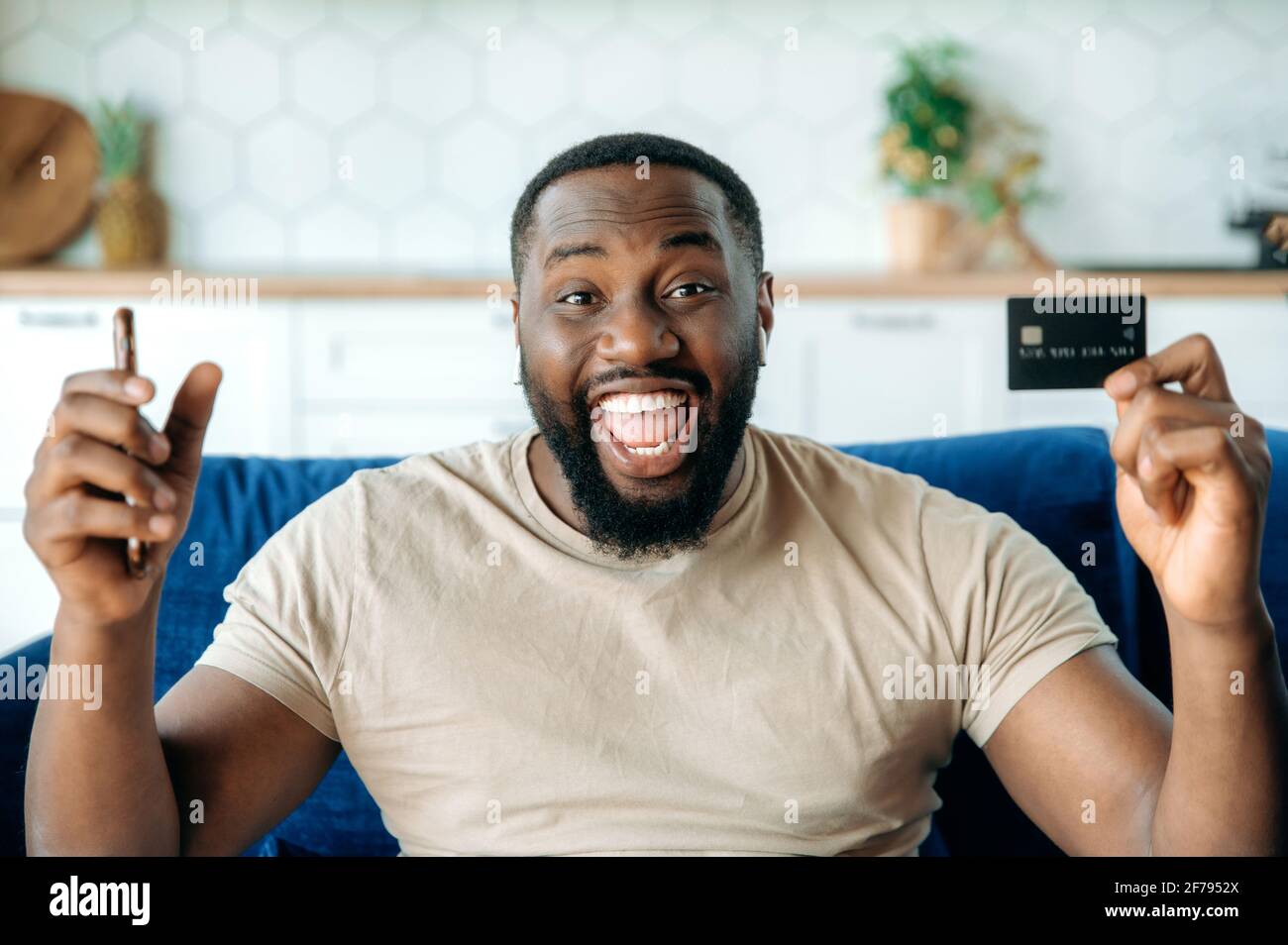 Joyeux joyeux jeune Africain américain barbu, dans des vêtements élégants, assis sur le canapé, tenant un smartphone et une carte de crédit, en faisant des gestes avec ses mains, souriant heureusement, a obtenu une réduction pour faire du shopping Banque D'Images