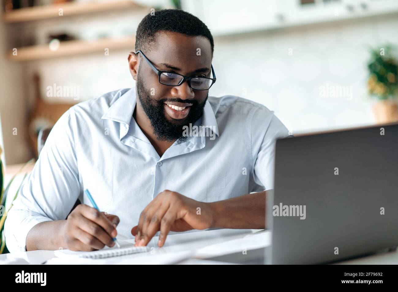 Sympathique et confiant, un homme afro-américain est barbu avec des lunettes, un courtier, un avocat ou un responsable travaillant à distance, prenant des notes, des conférences téléphoniques avec des collègues, apprenant à distance, souriant Banque D'Images