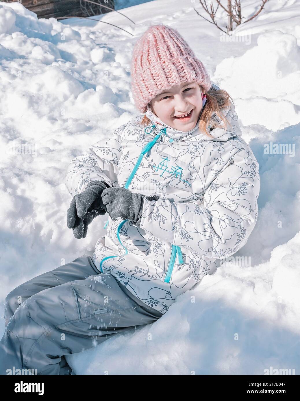 Jolie fille souriante dans des vêtements chauds est assise dans une banque de neige. Un enfant s'amuse en hiver. Origine ethnique caucasienne, enfance heureuse, activité en plein air. Pour Banque D'Images