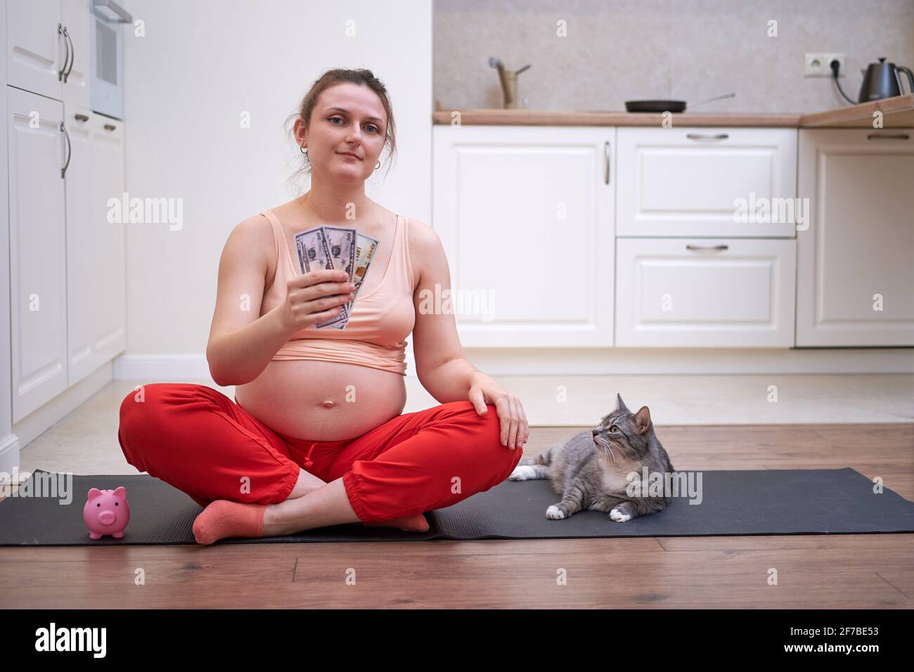 Jouer au sport pendant la grossesse et dépenser de l'argent. Femme enceinte faisant l'entraînement de forme physique dans la cuisine à la maison avec des dollars américains à la main Banque D'Images