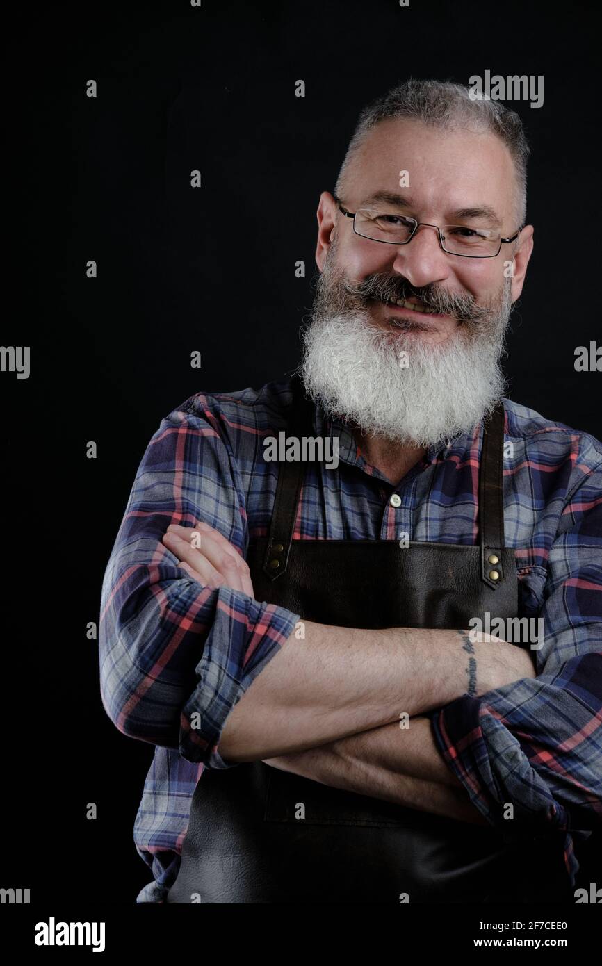 Portrait d'un beau homme mature barbu habillé tablier en cuir isolé sur fond noir, ouvrier caucasien avec barbe souriante Banque D'Images