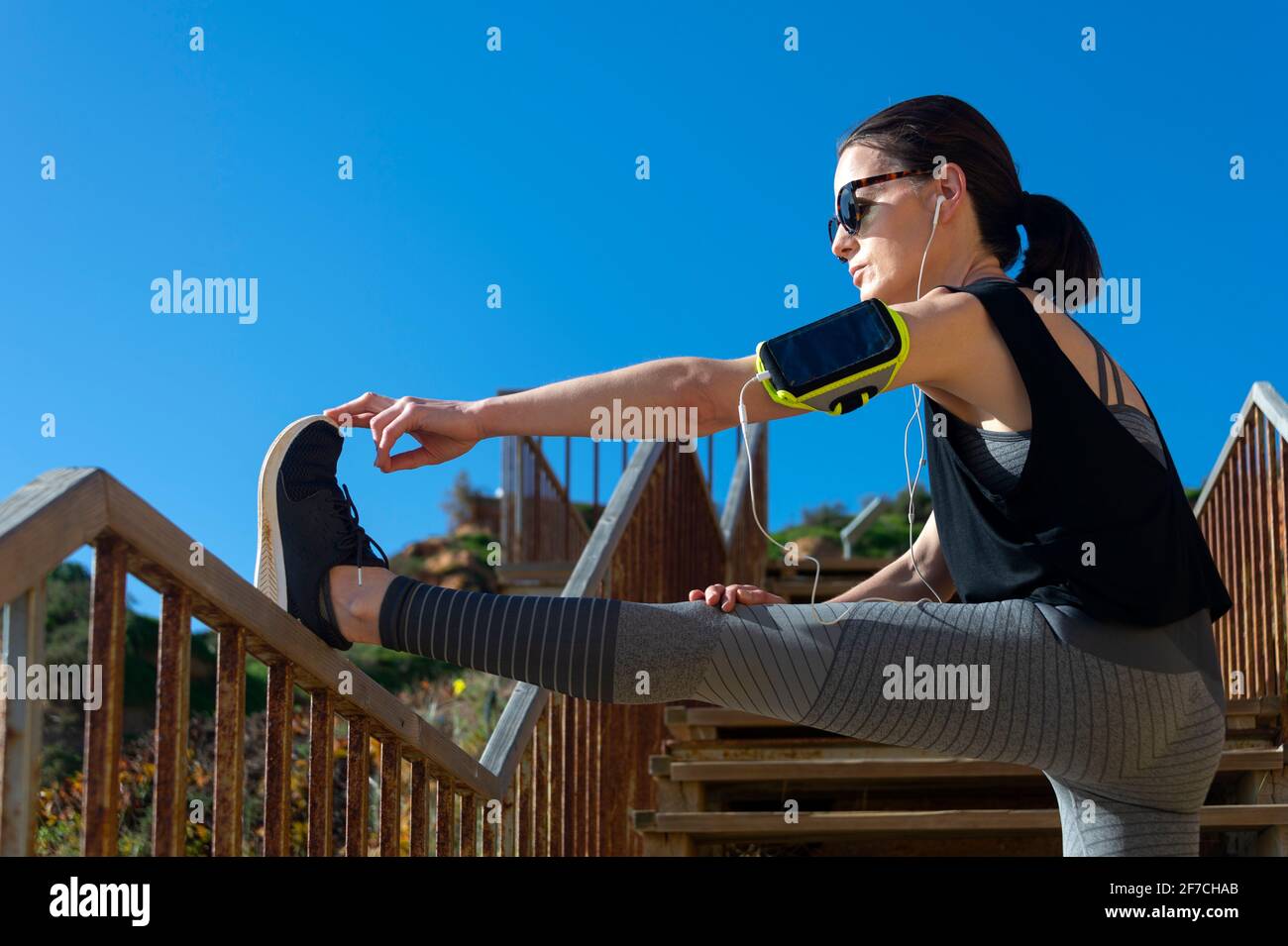 Femme athlétique étirant son hamstring, jambes entraînement fitness avant de s'entraîner à l'extérieur au soleil avec des écouteurs écoutant de la musique Banque D'Images