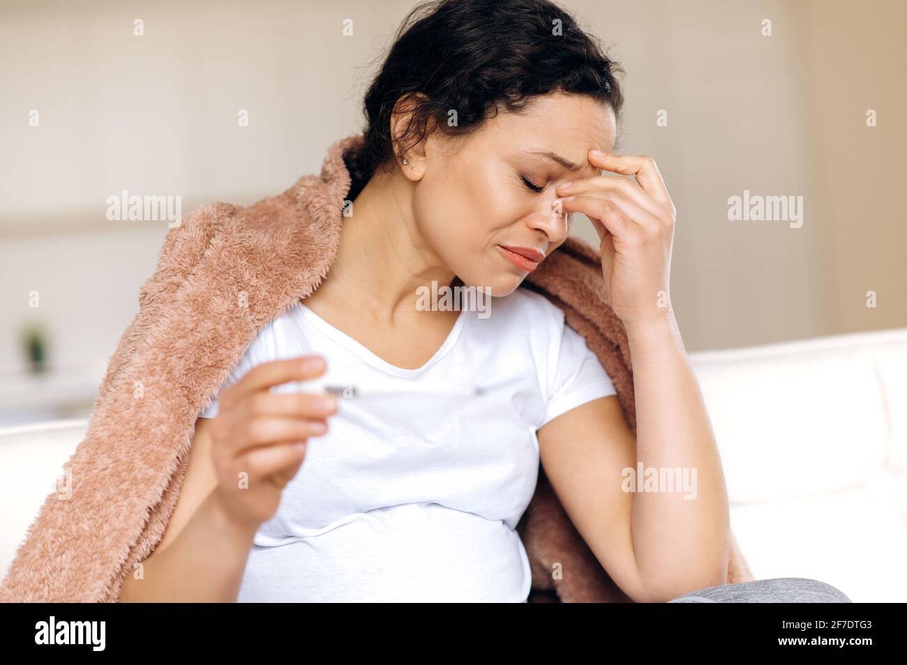 Femme enceinte de race mixte malade exténuée assise au salon sur le canapé sous la couverture, masser ses yeux avec la main tout en prenant la température avec le thermomètre, elle a un rhume, la grippe Banque D'Images