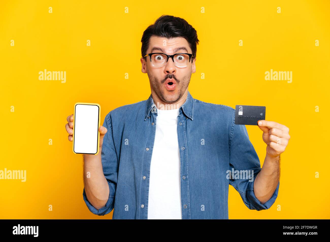 Stupéfait choqué caucasien élégamment habillé gars avec des lunettes, tient un smartphone et une carte de crédit dans les mains, regarde surpris à la caméra, se tenir sur un arrière-plan isolé orange, e-banking concept Banque D'Images