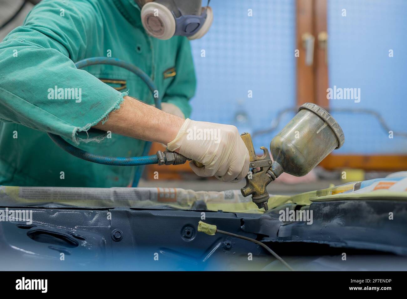 Homme en combinaison verte préparant un compartiment moteur d'une voiture pour une dernière couche de peinture. Homme pulvérisant du mastic liquide gris ou du stuc sur les panneaux de voiture en dernier Banque D'Images
