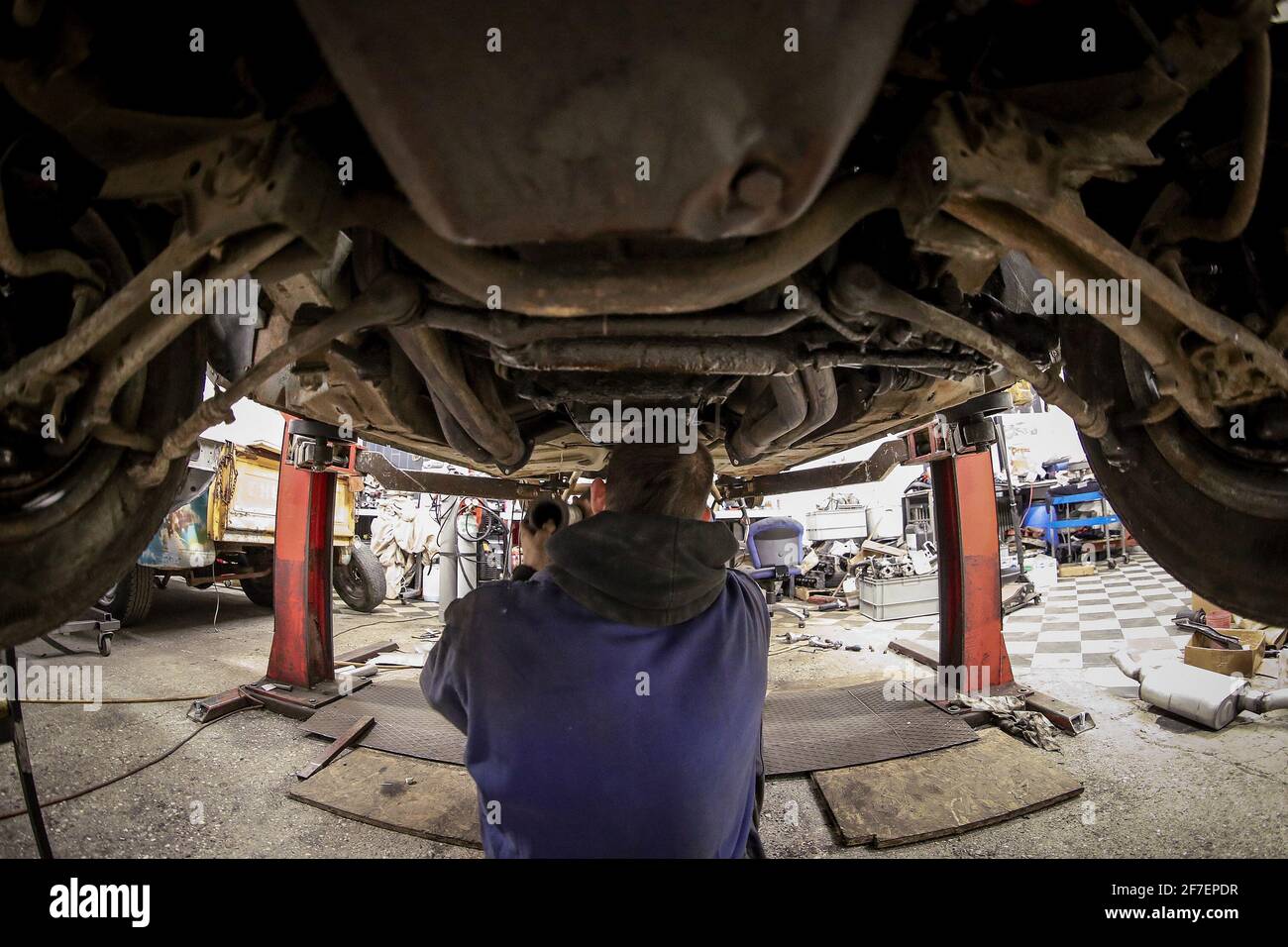 Un homme avec une coupe de cheveux mohawk inspecte le dessous d'une vieille voiture rouillée sur un ascenseur et fixe l'échappement. Sous la voiture dans un atelier. Banque D'Images