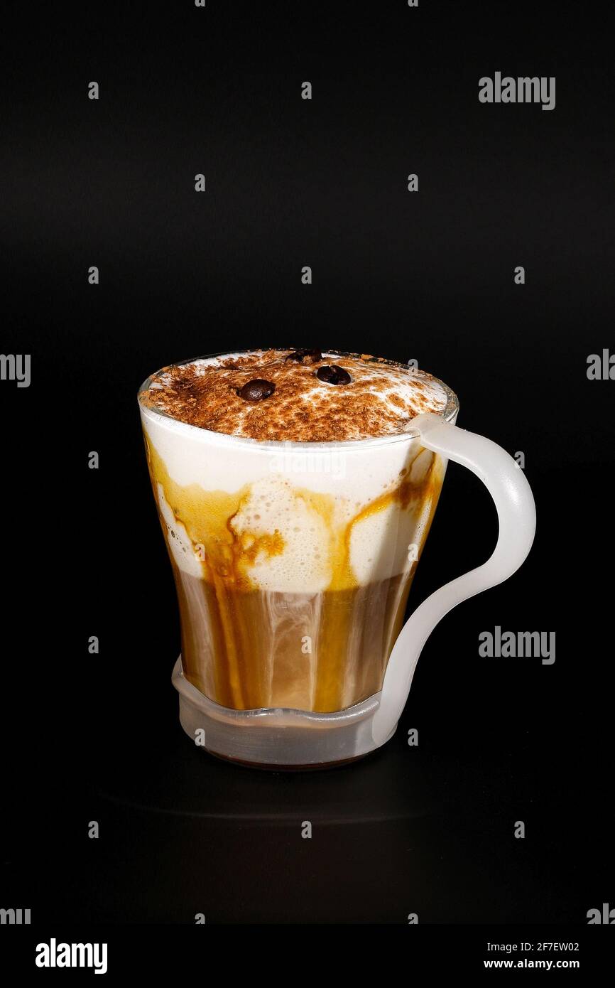 Chocolat chaud ou boisson au café dans une tasse en verre avec support, isolée sur le noir. Chapelure de noix de coco visible, lait, crème fouettée et autres ingrédients sucrés. Banque D'Images