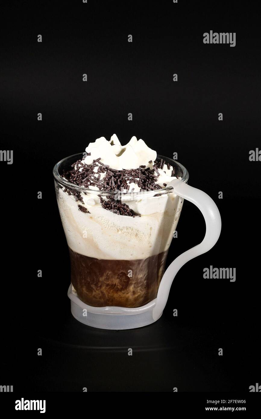 Chocolat chaud ou boisson au café dans une tasse en verre avec support, isolée sur le noir. Chapelure de noix de coco visible, lait, crème fouettée et autres ingrédients sucrés. Banque D'Images