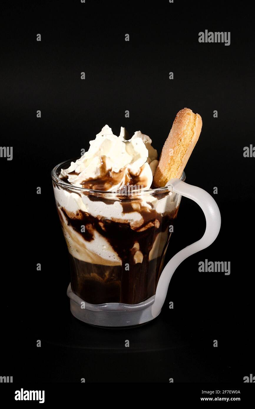 Chocolat chaud ou boisson au café dans une tasse en verre avec support, isolée sur le noir. Chapelure de noix de coco visible, lait, crème fouettée et autres ingrédients sucrés Banque D'Images