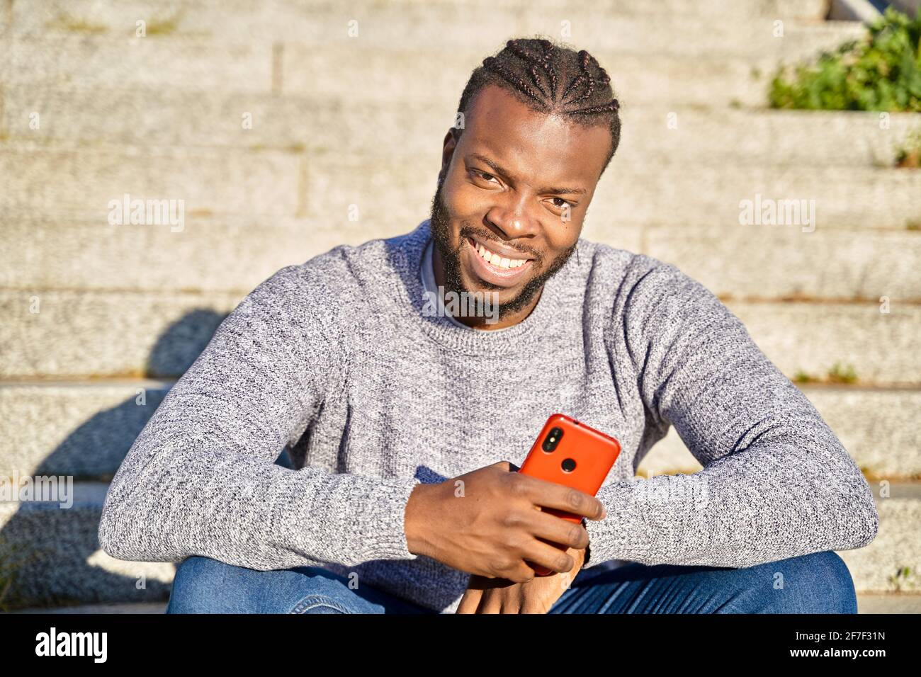 Gros plan d'un homme afro-américain assis dans les escaliers souriant, regardant la caméra, par une journée ensoleillée. Joyeux jeune Latino homme. Photo de haute qualité Banque D'Images