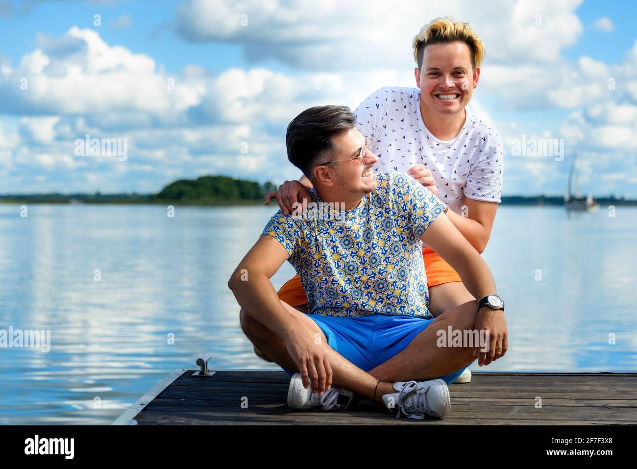 joyeux couple gay souriant assis sur une jetée en bois contre lac et ciel bleu Banque D'Images