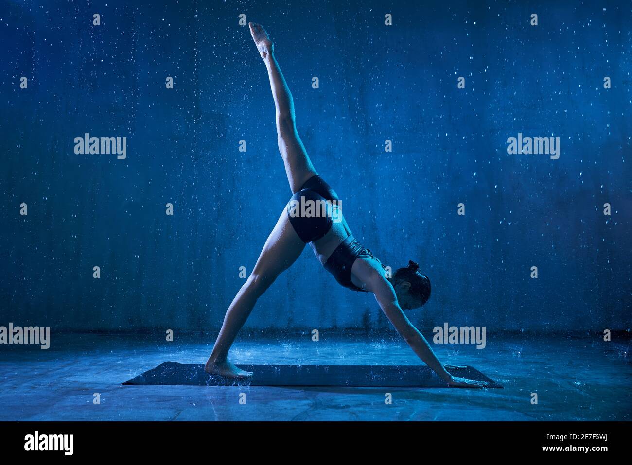 Femme sportive attrayante portant des vêtements de sport noirs debout dans l'asana de yoga avec une jambe relevée sous la pluie. Forte flexible fille debout dans le bas face chien, adho mukha svanasana pose. Banque D'Images