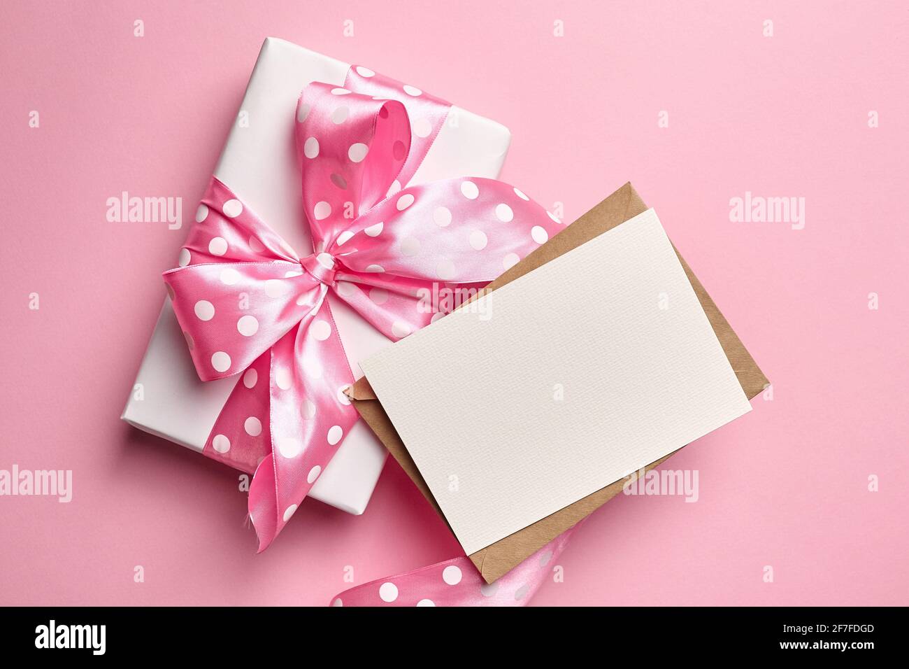 Maquette de carte de vœux avec boîte cadeau ornée d'un noeud sur fond de papier rose. Carte de vœux blanche ou invitation et ruban à pois roses, vue du dessus Banque D'Images
