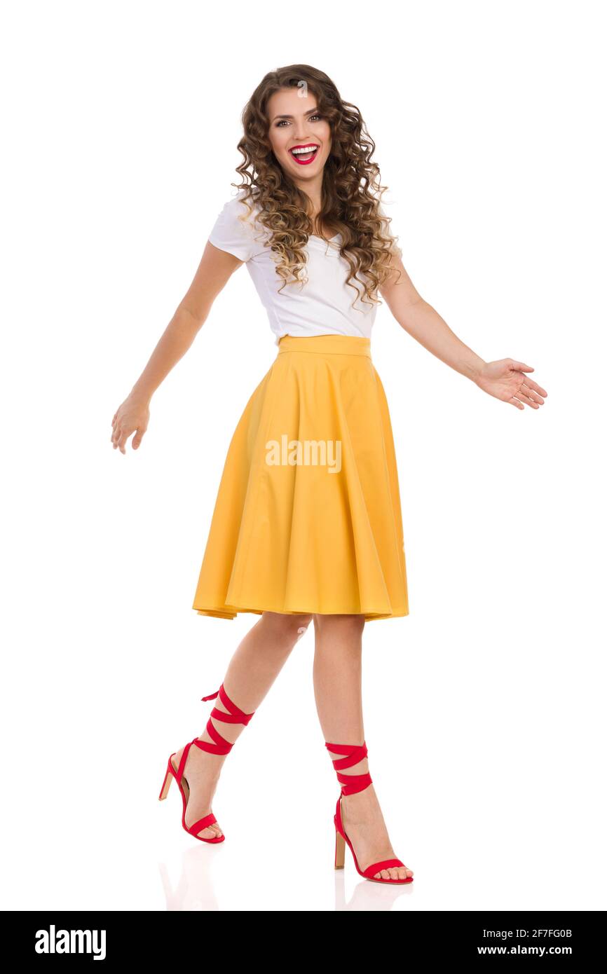 Belle jeune femme en haut blanc, jupe jaune et talons hauts rouges est marche et regarde le camer et rire. Vue latérale. Plein studio tourné i Banque D'Images