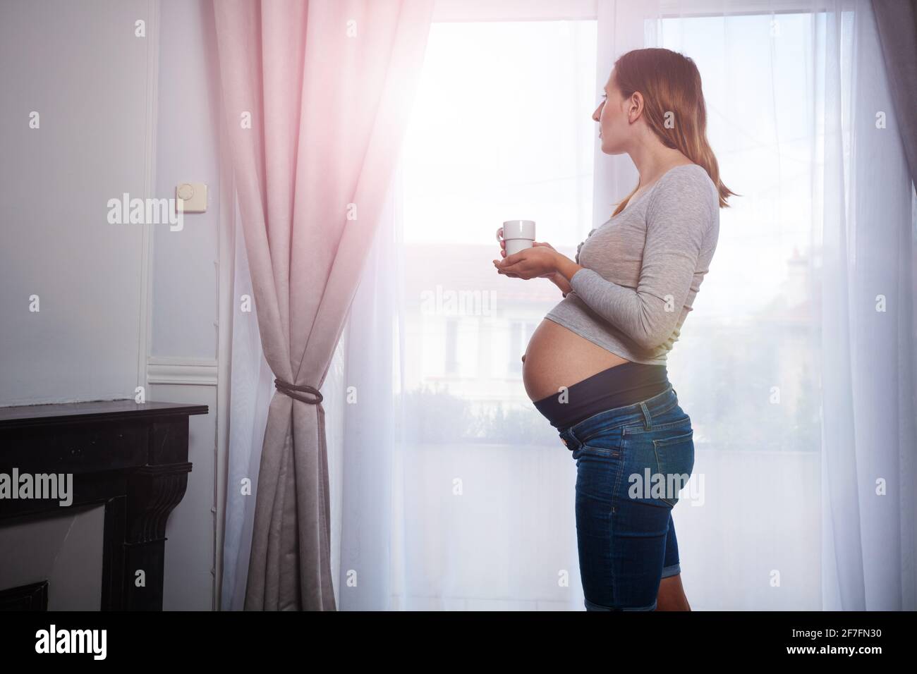 Femme se tenir près de la fenêtre, boire du thé en regardant dehors Banque D'Images