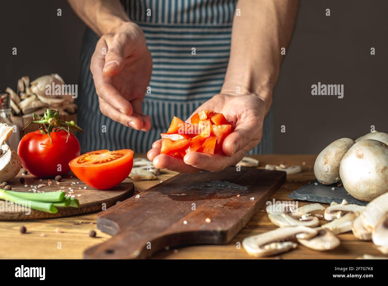 Un chef masculin dans un tablier bleu coupe des tomates fraîches appétissantes avec un couteau pour préparer un plat. Concept du processus de cuisson. Banque D'Images